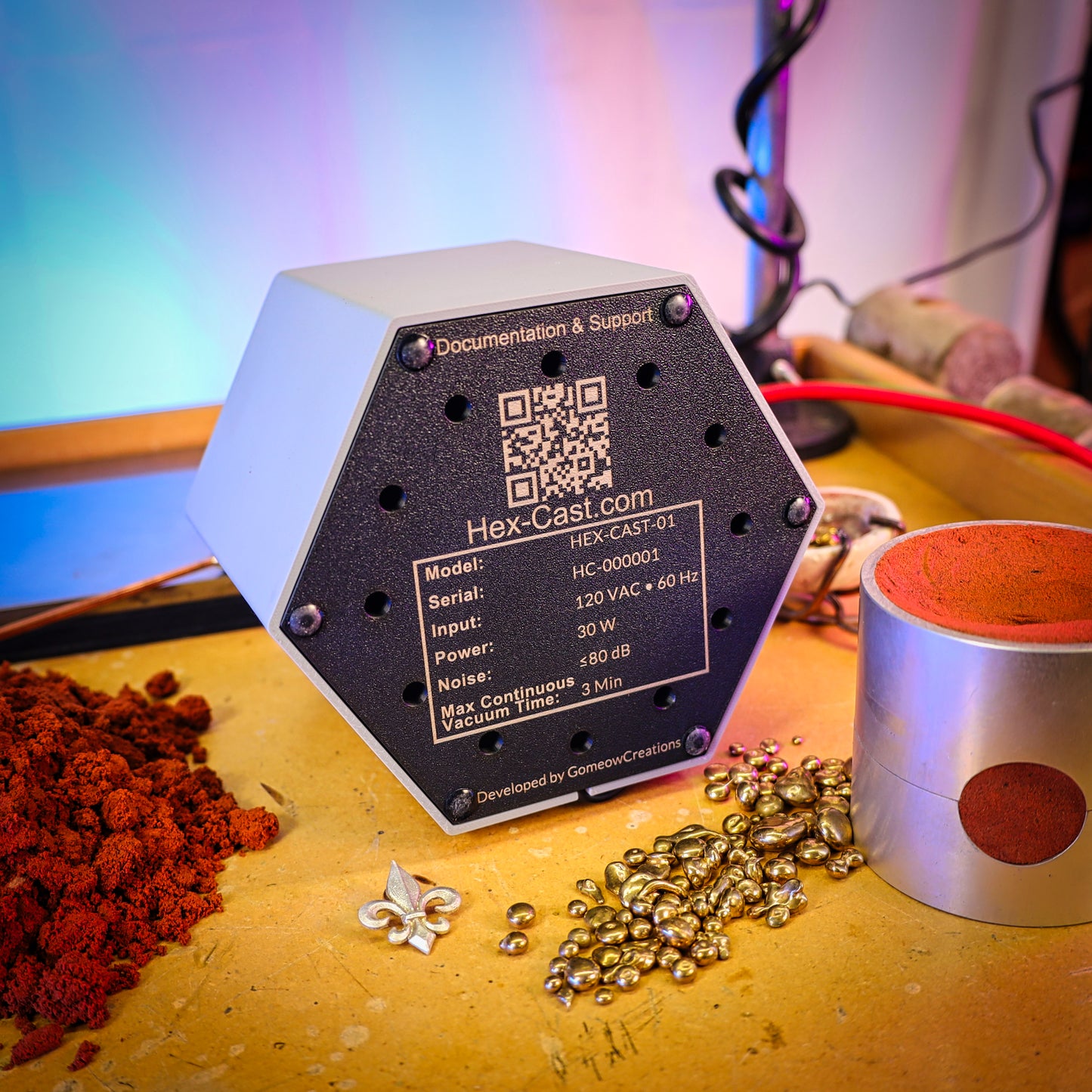 A Hex-Cast sitting on a workbench surrounded by red casting sand, brass casting grain, and a metal casting flask, with a colorful blurred background.