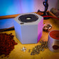 A  Hex-Cast jewelry casting machine sits on a wooden workbench surrounded by red casting sand, metallic beads, and a small silver pendant, with colorful ambient lighting in the background.