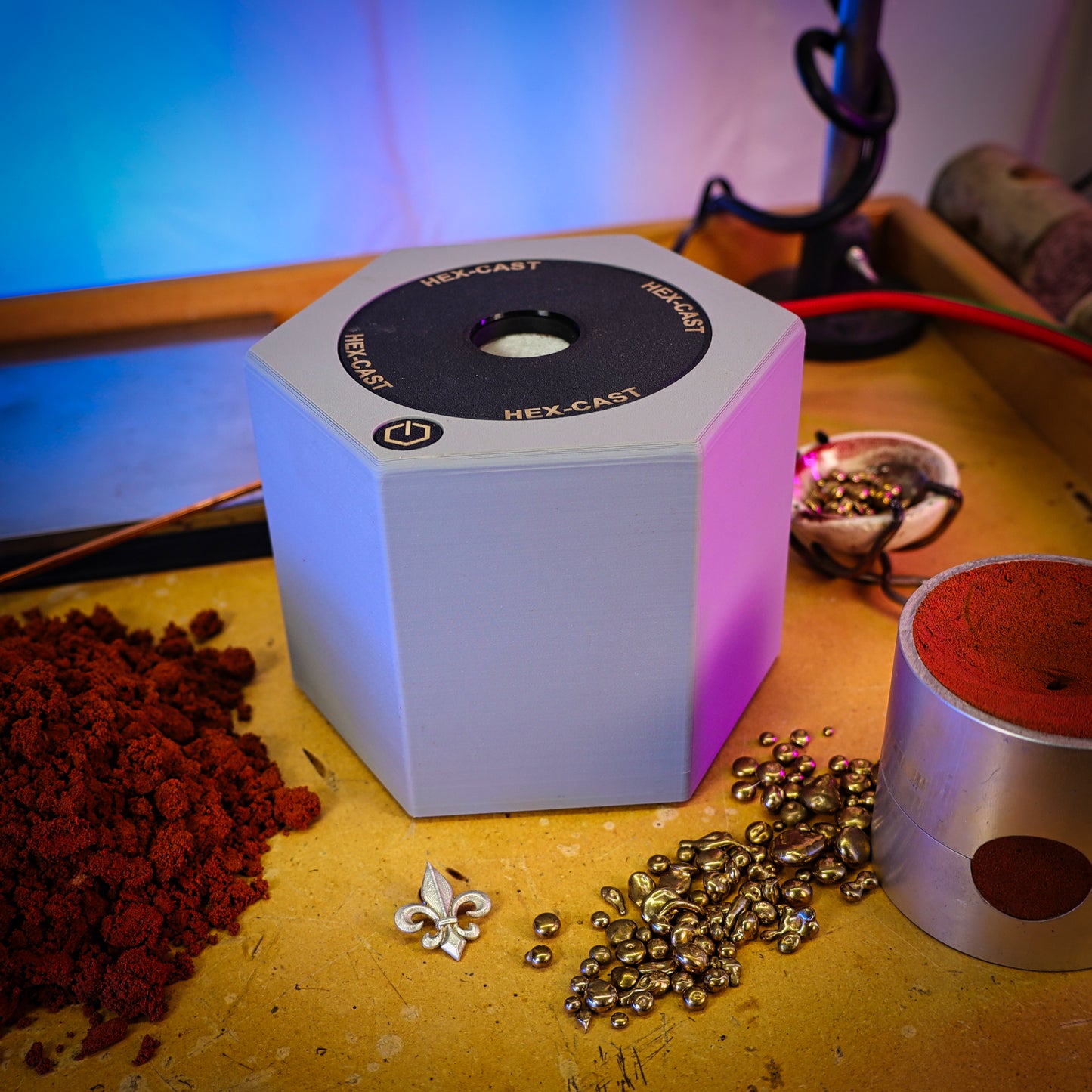 A  Hex-Cast jewelry casting machine sits on a wooden workbench surrounded by red casting sand, metallic beads, and a small silver pendant, with colorful ambient lighting in the background.