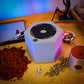 A  Hex-Cast jewelry casting machine sits on a wooden workbench surrounded by red casting sand, metallic beads, and a small silver pendant, with colorful ambient lighting in the background.