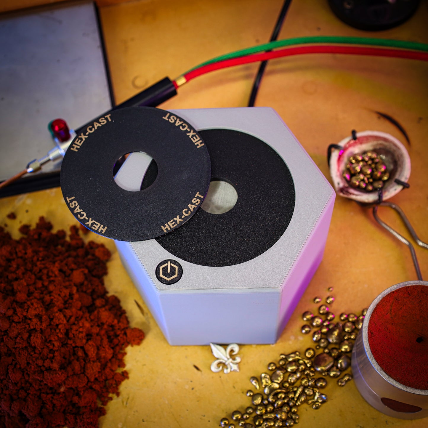 A hexagonal metal casting mold or mold box with a black circular insert labeled HEX-CAST, shown on a workbench surrounded by metal beads, red powder, and casting tools.