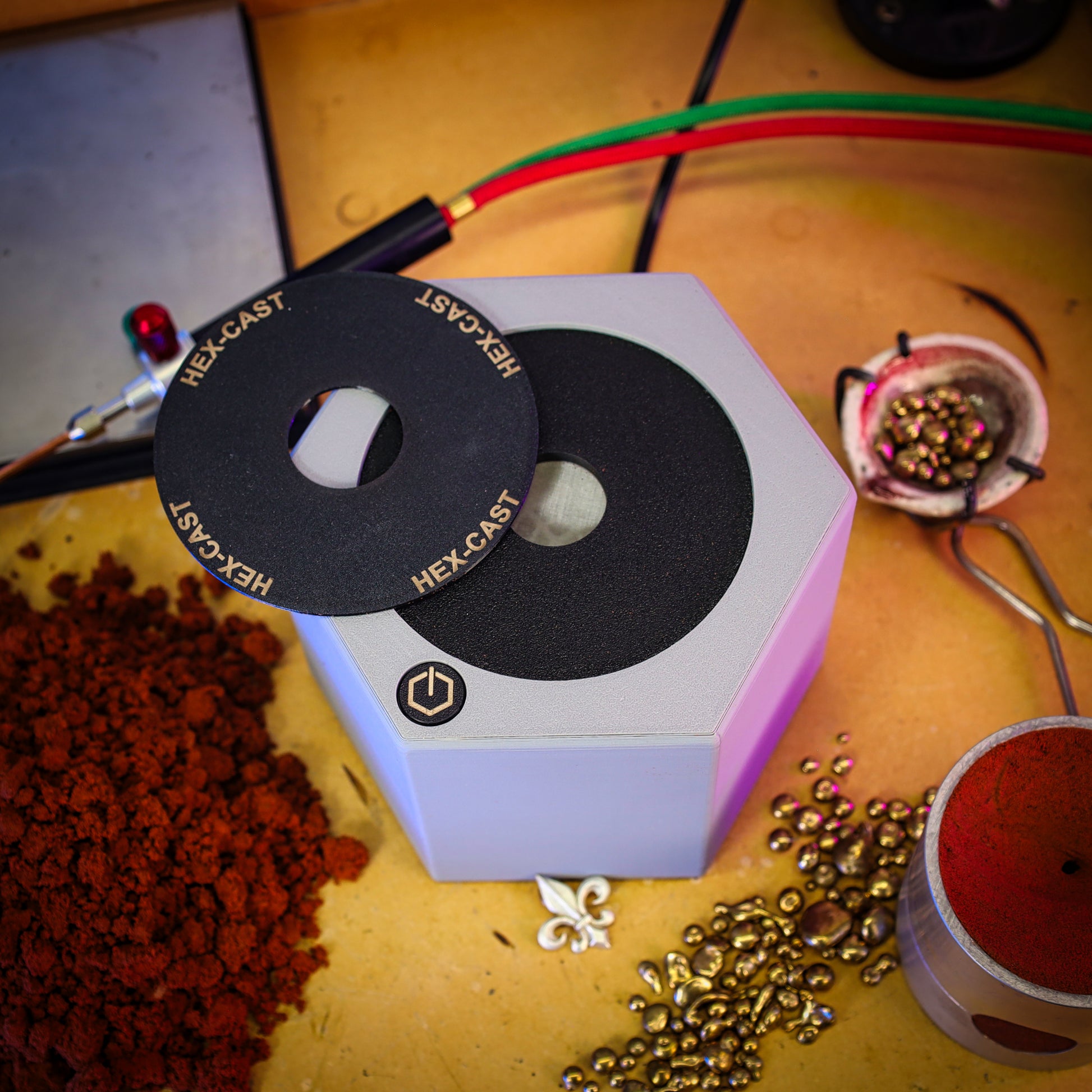 A hexagonal metal casting mold or mold box with a black circular insert labeled HEX-CAST, shown on a workbench surrounded by metal beads, red powder, and casting tools.